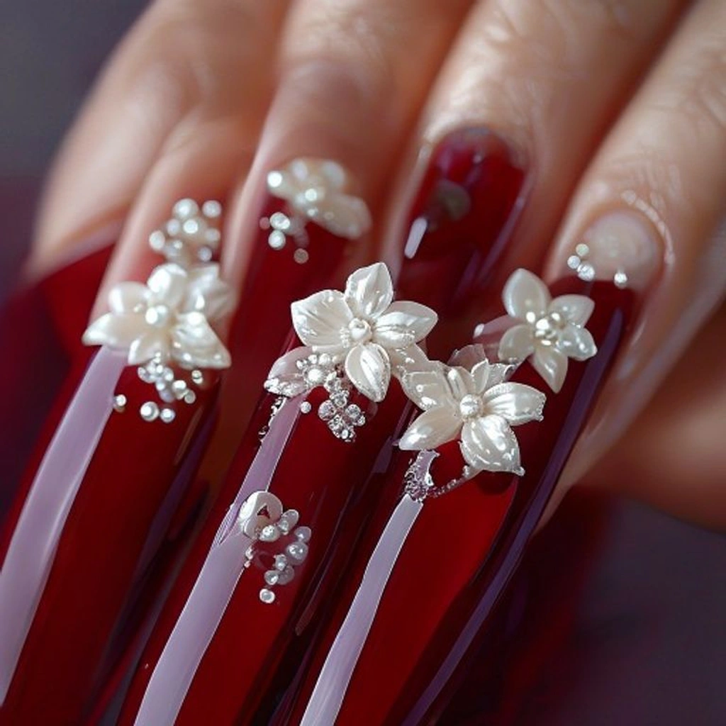 Close-up of long stiletto nails in a deep crimson gel base, adorned with prominent 3D white acrylic flower embellishments and small pearls in the center of the flowers. High-glamour, luxury studio lighting.
